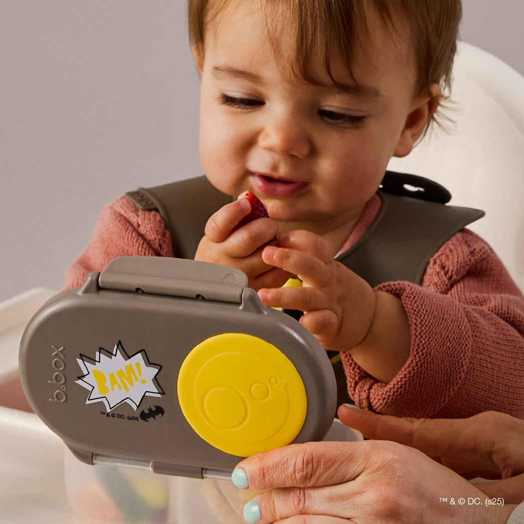 Child using a b.box snack container with a yellow lid, sitting in a high chair.