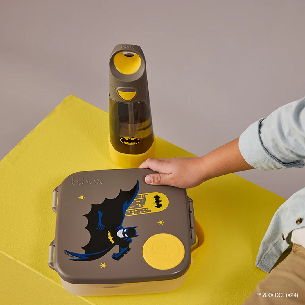 Child's hand opening a Batman-themed bento box on a yellow surface with a bottle in the background.