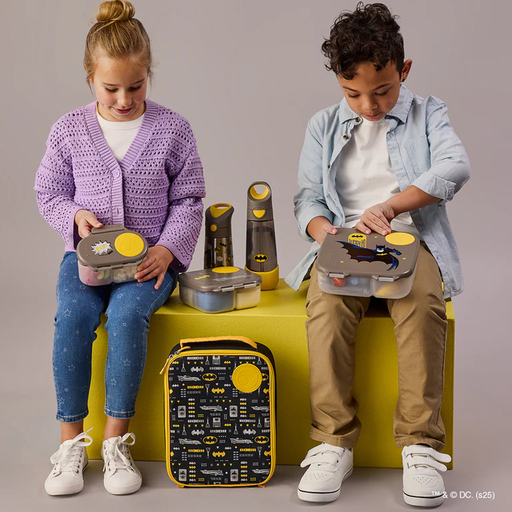 Two children playing with Batman-themed lunch boxes on a yellow surface.