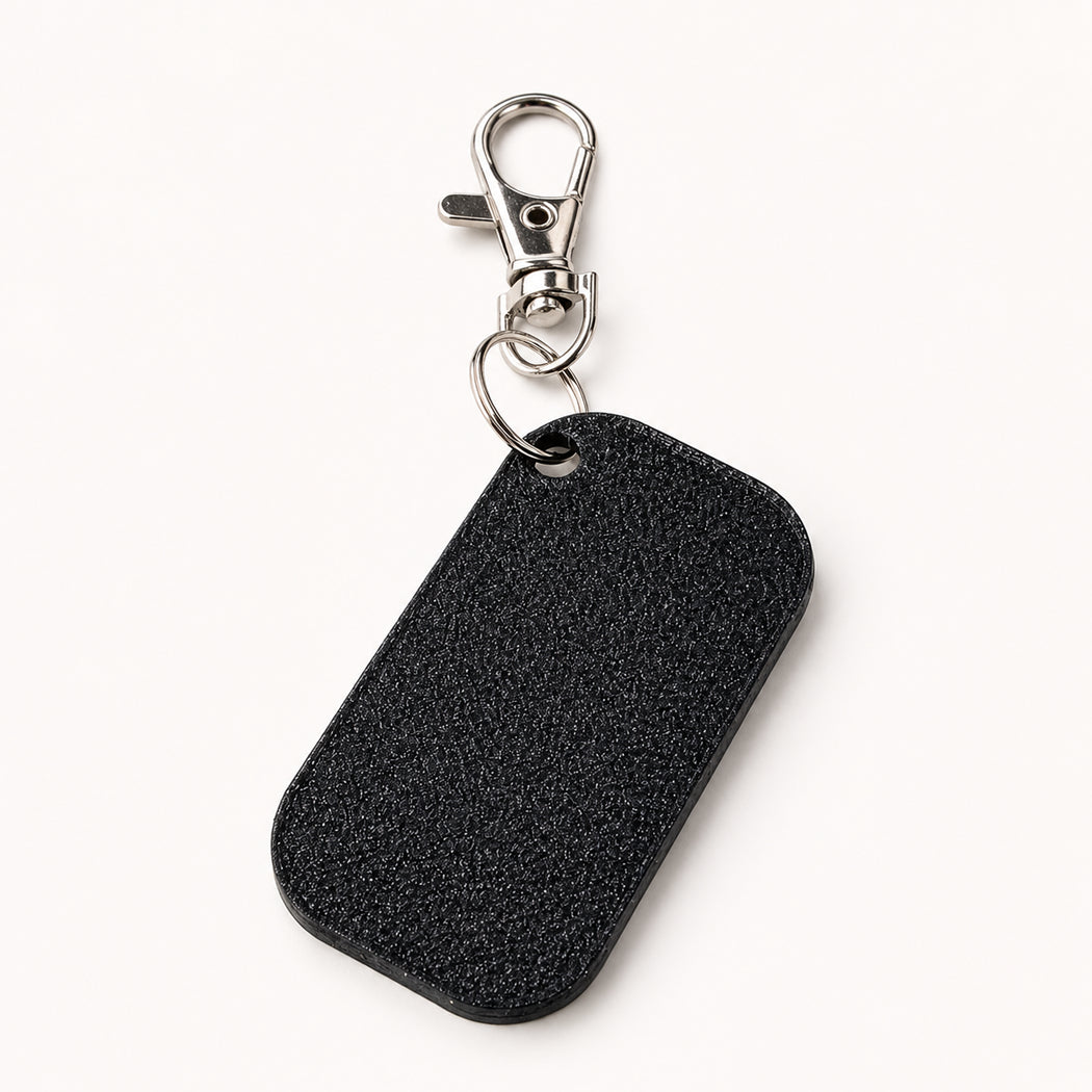 A black textured leather dog tag keychain with a silver-tone lobster clasp, shown on a clean off-white background.