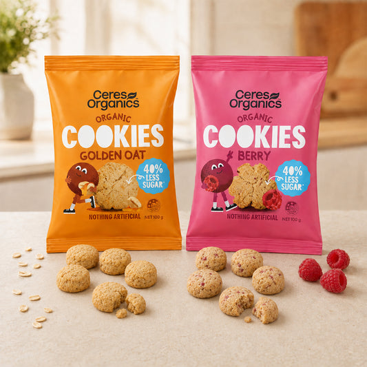 Two bags of Ceres Organics Organic Cookies, one golden oat flavor in orange and one berry flavor in pink, displayed on a light beige surface with scattered cookies and fresh raspberries.
