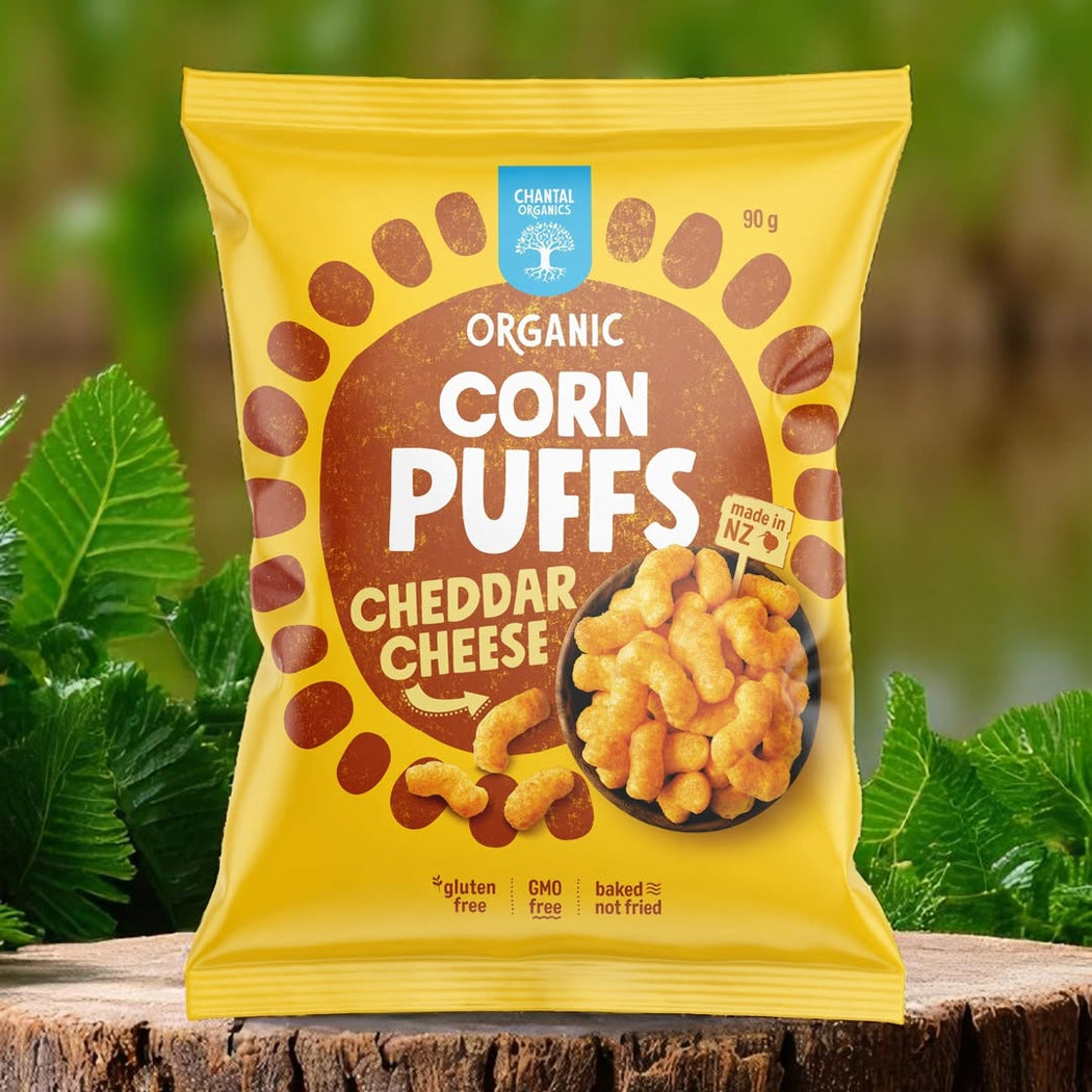Packaging of organic corn puffs with cheddar cheese flavor on a wooden surface with green leaves in the background.