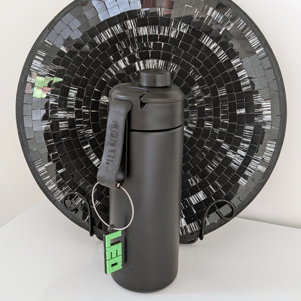 Black water bottle with a keychain attached, placed in front of a decorative black and white mosaic wall.