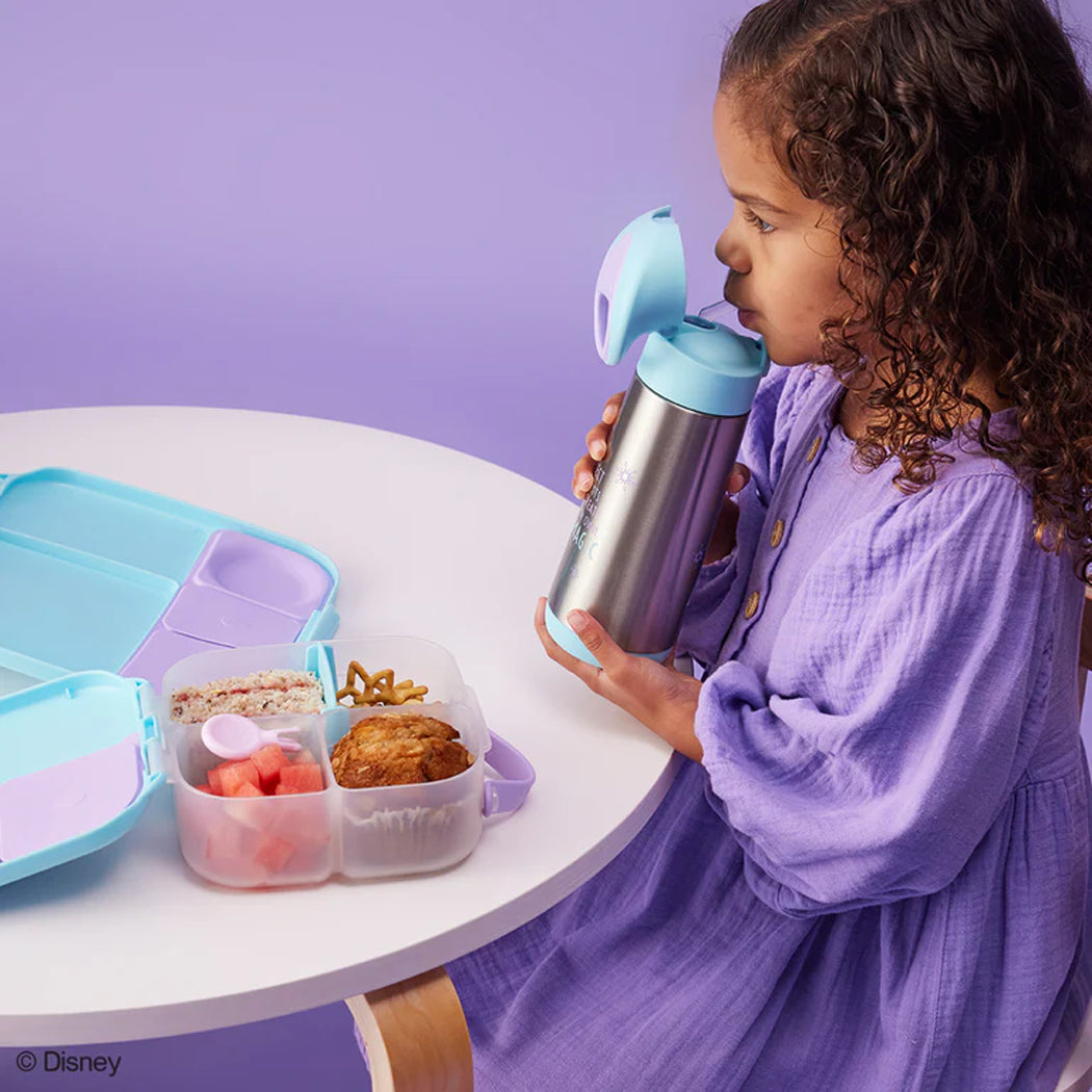Child drinking from a thermos with a purple table and lunchbox in the background