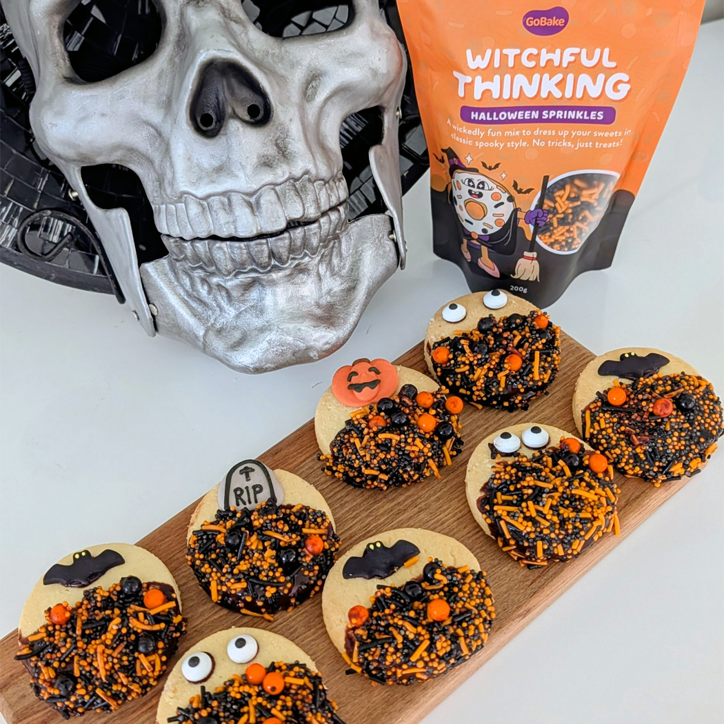 Halloween-themed cookies on a wooden board with a skull and 'Witchful Thinking' sprinkles packaging in the background.