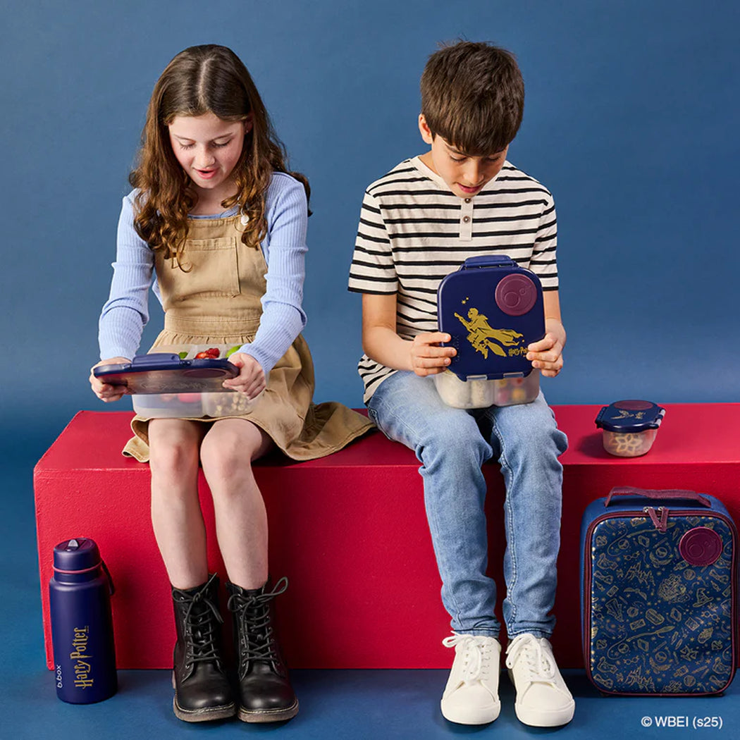 Two children sitting on a red bench with lunch boxes against a blue background