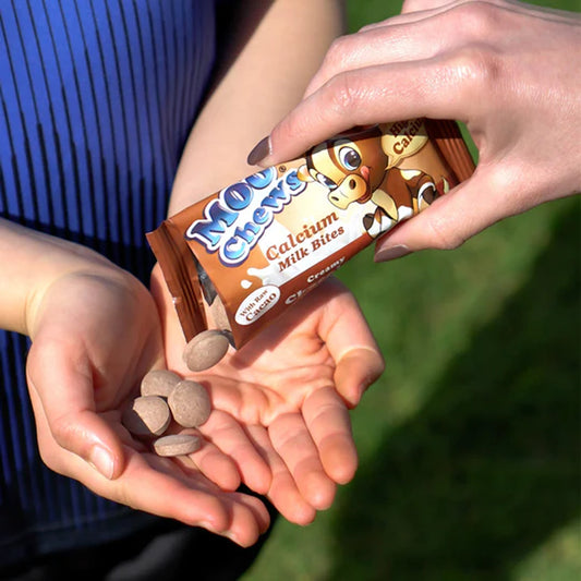Two hands holding a package of 'Moo Chews Calcium Milk Bites' with stones in the palm against a blurred green background.