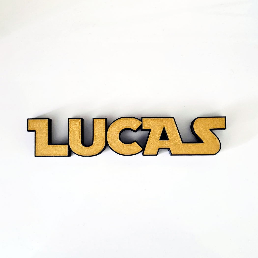 A 3D wooden name sign spelling 'LUCAS' in a bold, stylized font, featuring a natural light wood finish with black edges, set against a plain white background.