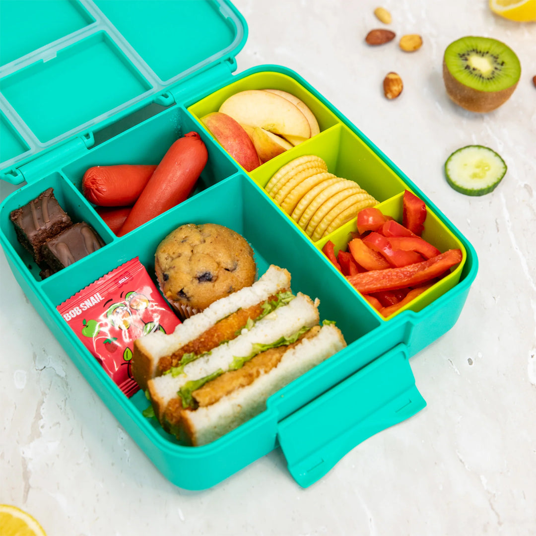 Colorful bento box with various snacks including sandwiches, cookies, and fruits on a light background.