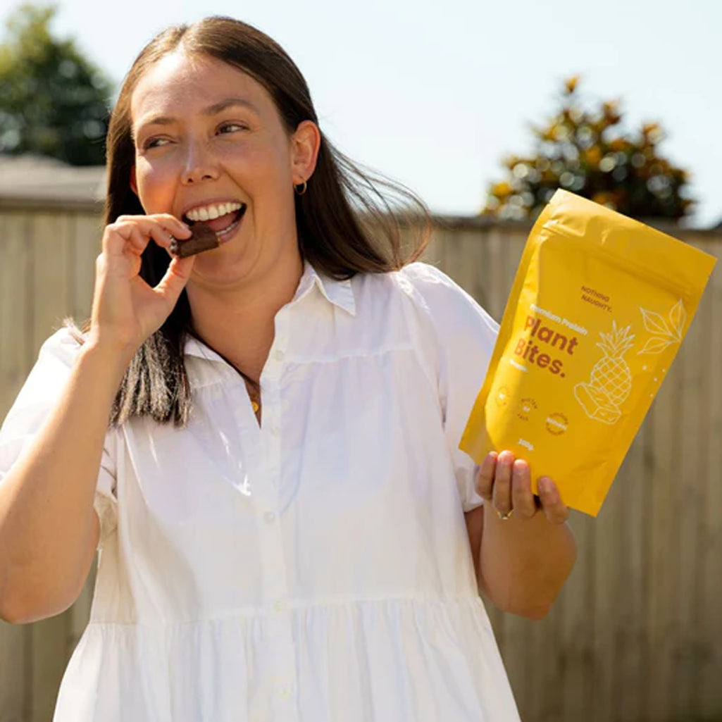 Woman eating a snack from a yellow 'Plant Bites' package outdoors.