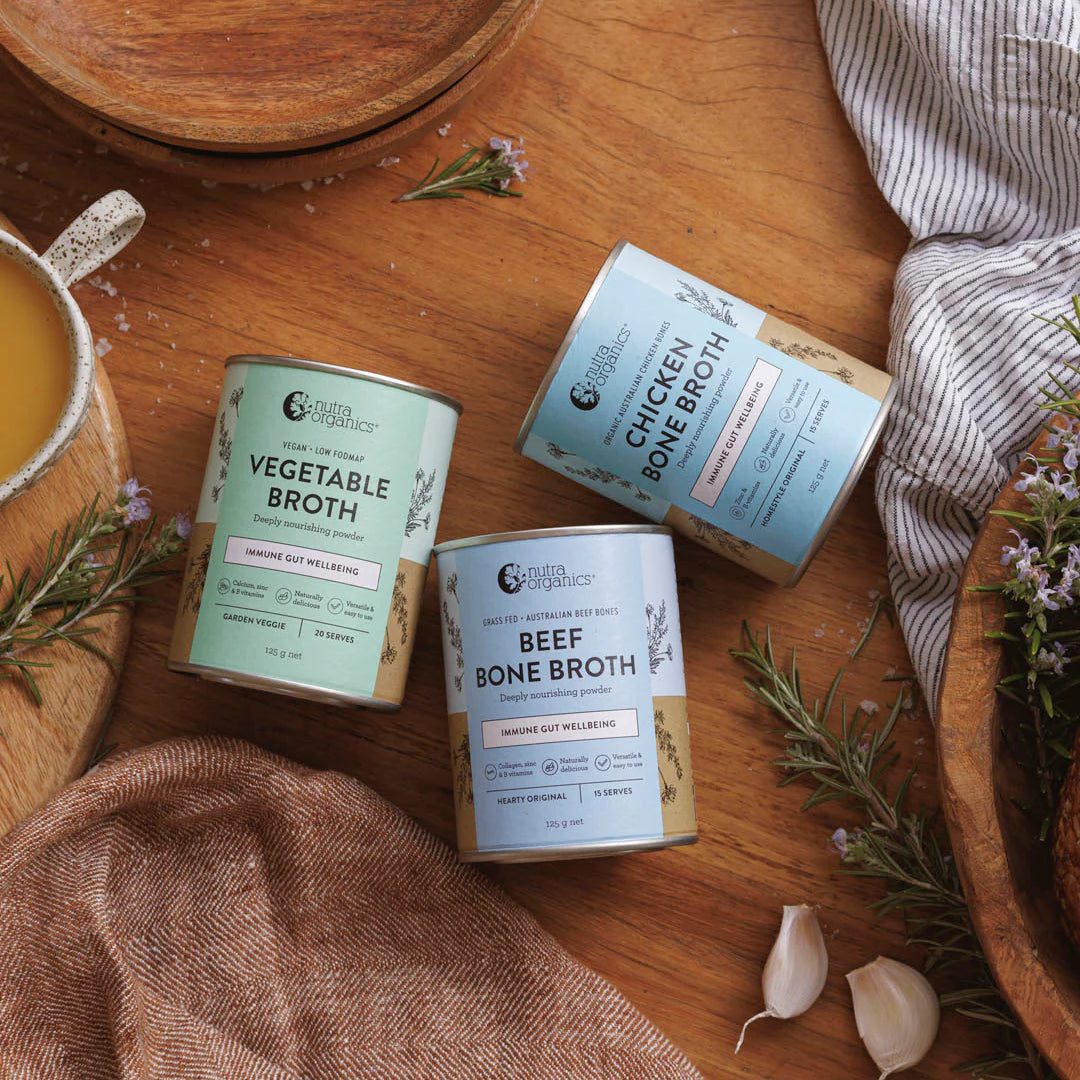 Three cans of bone broth on a wooden table with herbs and a mug.
