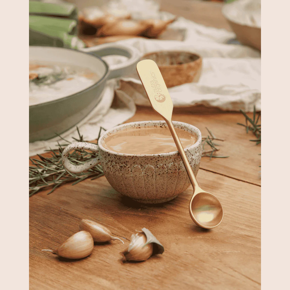 Cup of soup with a spoon on a wooden table, surrounded by garlic and rosemary.