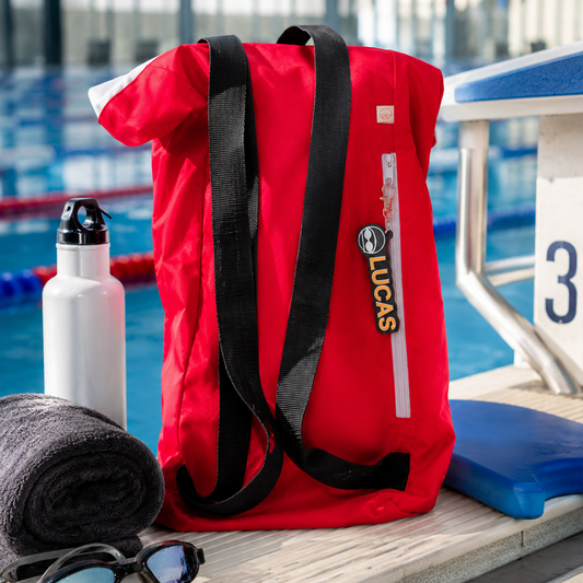 A red roll-top swim bag with black straps and a vertical white zipper pull labeled LUCAS, placed on a poolside deck with a white bottle, towel, and swim goggles nearby.