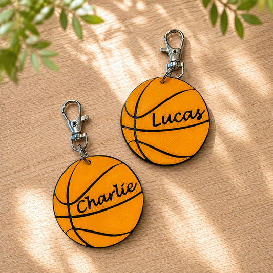 Two personalized basketball-shaped keychains with silver clasp hardware, displayed on a light wood surface with soft leaf shadows. One keychain is labeled 'Charlie' and the other 'Lucas'.