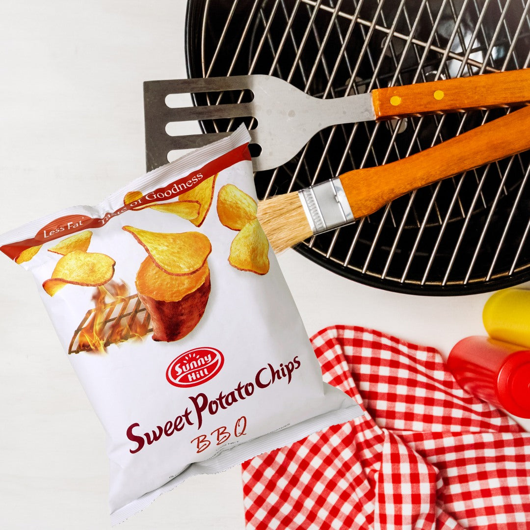 Bag of Sunny Delight Sweet Potato Chips BBQ next to a grill with utensils and a checkered cloth.