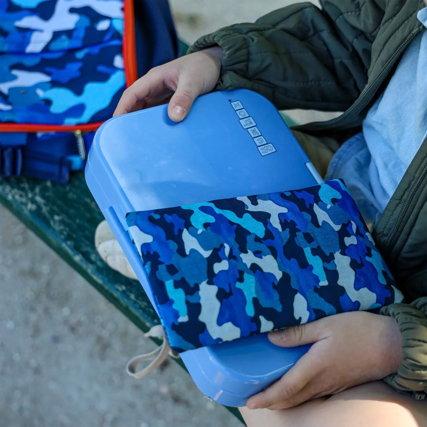 Person holding a blue lunch box with camouflage pattern on a bench