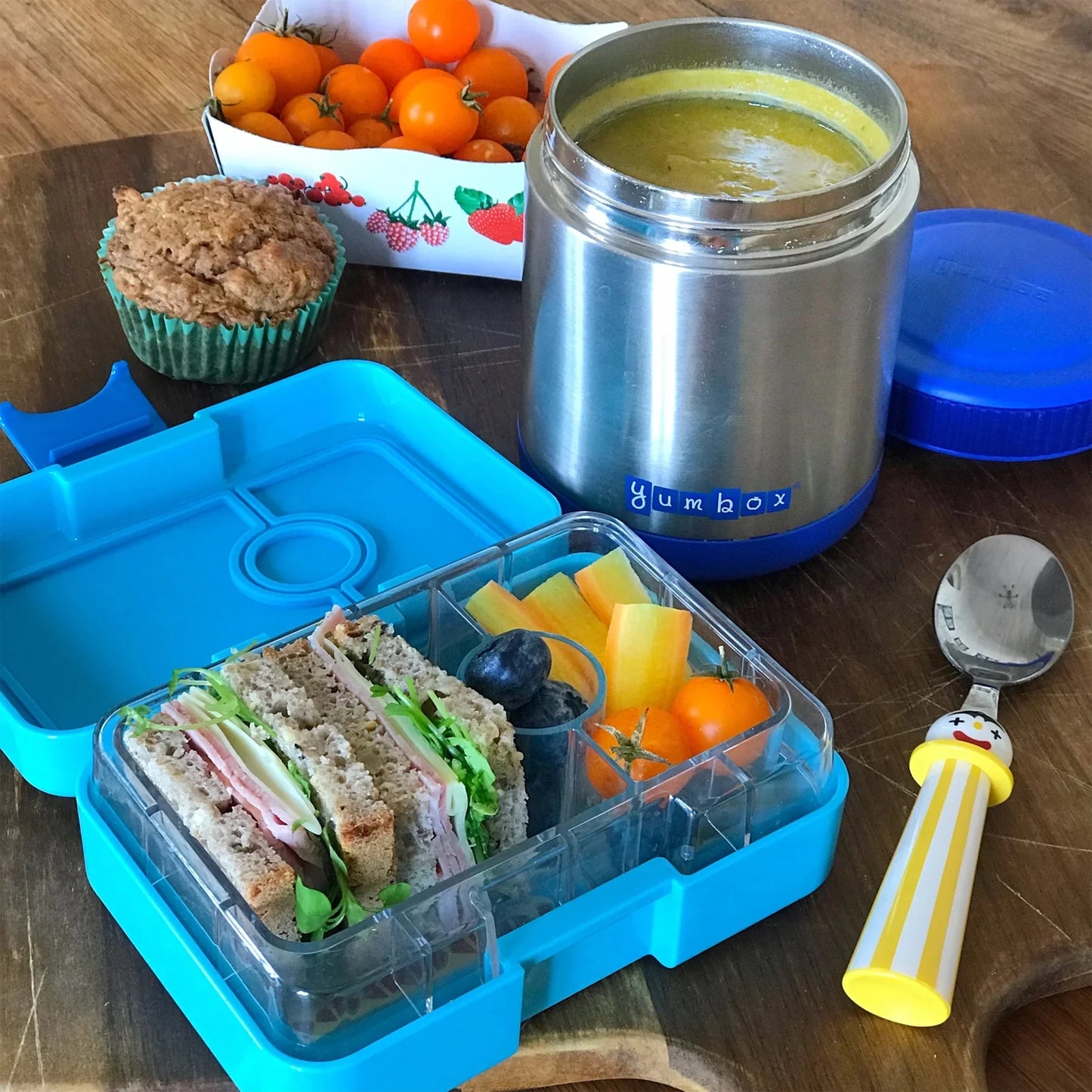 Lunchbox with sandwich, fruit, and yogurt, accompanied by a thermos and muffin on a wooden table.