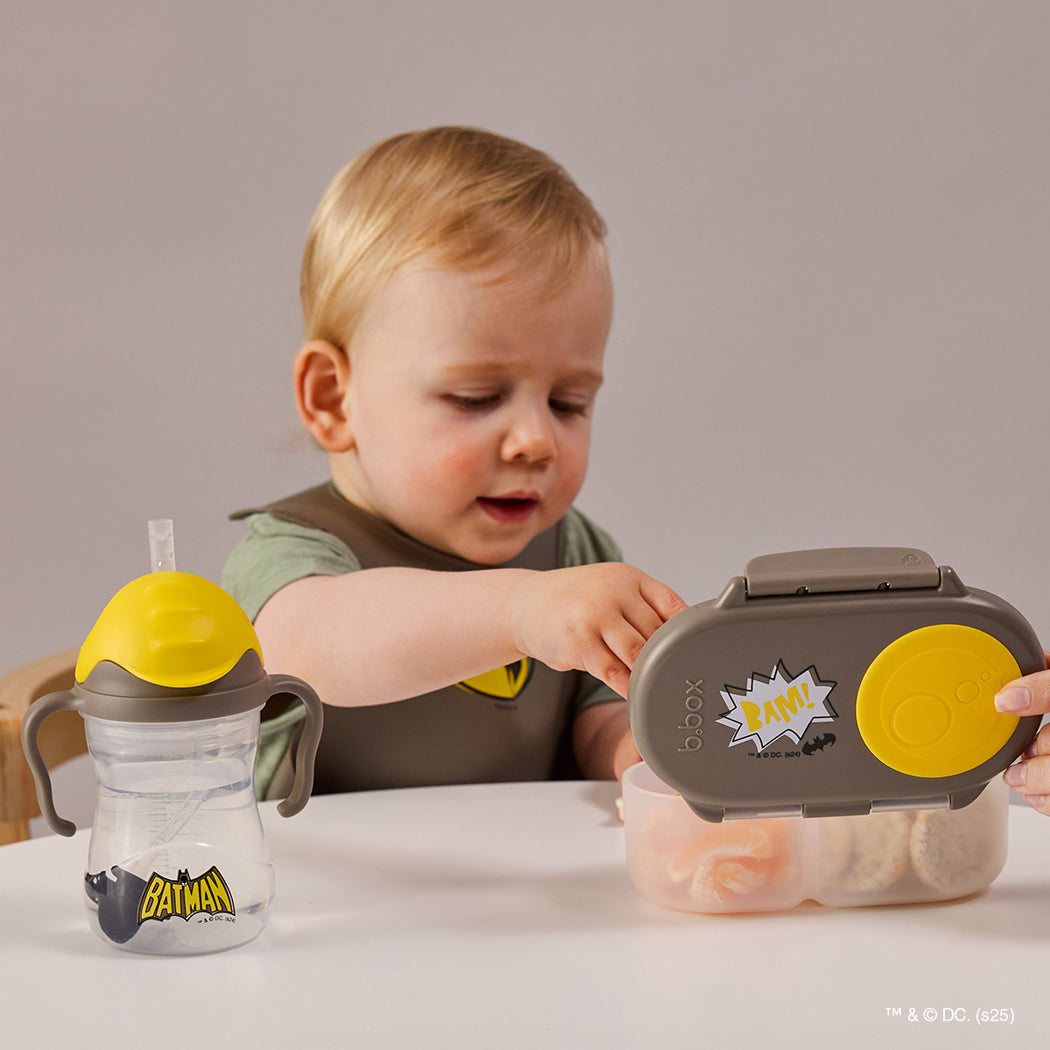Child playing with a Batman-themed snack container and sippy cup on a table.