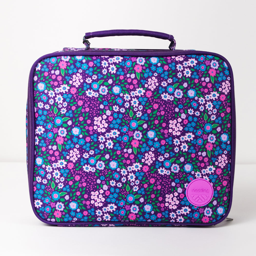 A rectangular lunch bag with a purple top handle and trim, featuring an all-over floral print in purple, blue, pink, and white. A pink circular 'gourmet' brand patch is visible on the lower right front.