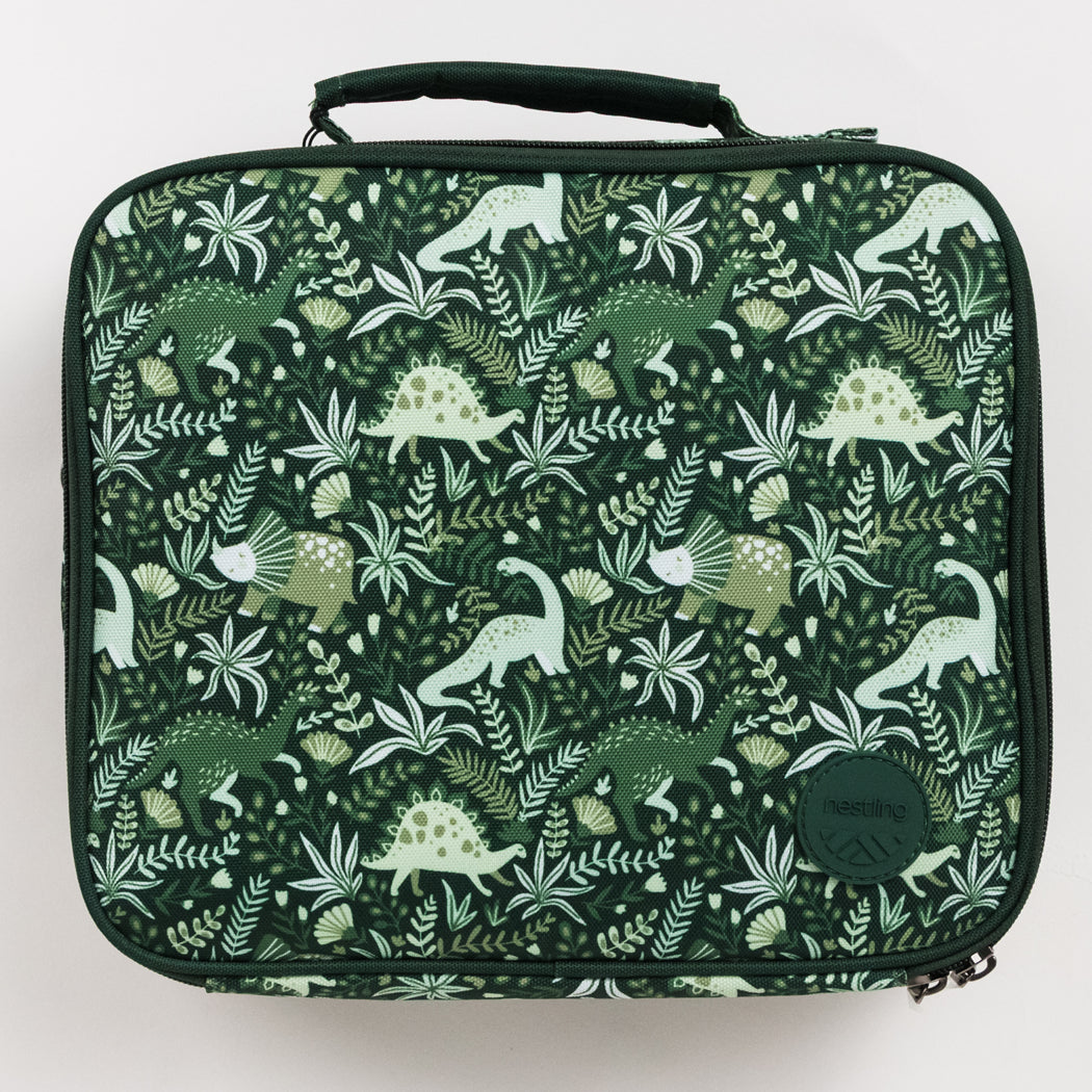A square insulated lunch bag with a dark green background and an all-over dinosaur and foliage print in various shades of green and off-white. The bag features a top carry handle, a zippered closure, and a circular brand patch on the bottom right.