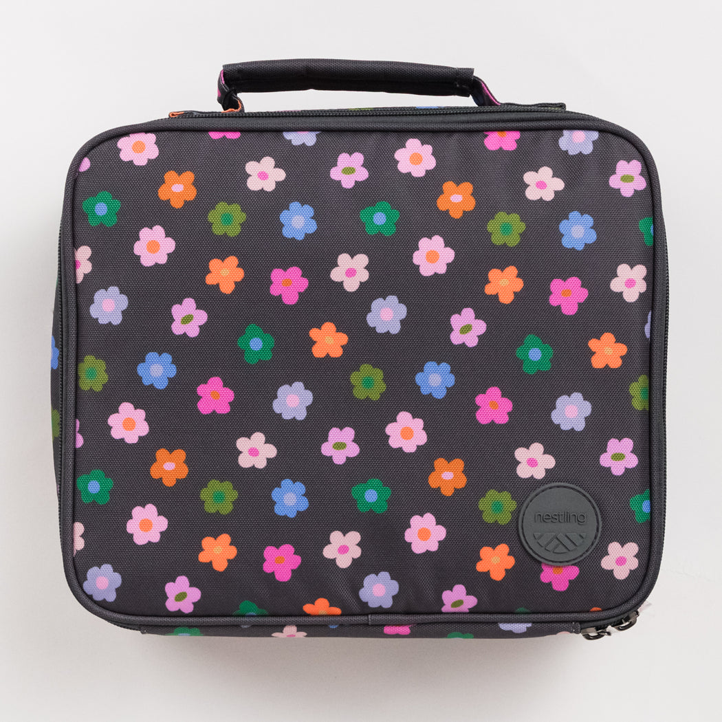 A rectangular insulated lunch bag with a black handle, featuring a dark gray body covered in a colorful pattern of small flowers in pink, orange, green, blue, and purple. A circular black logo patch for 'nestling' is visible on the bottom right.