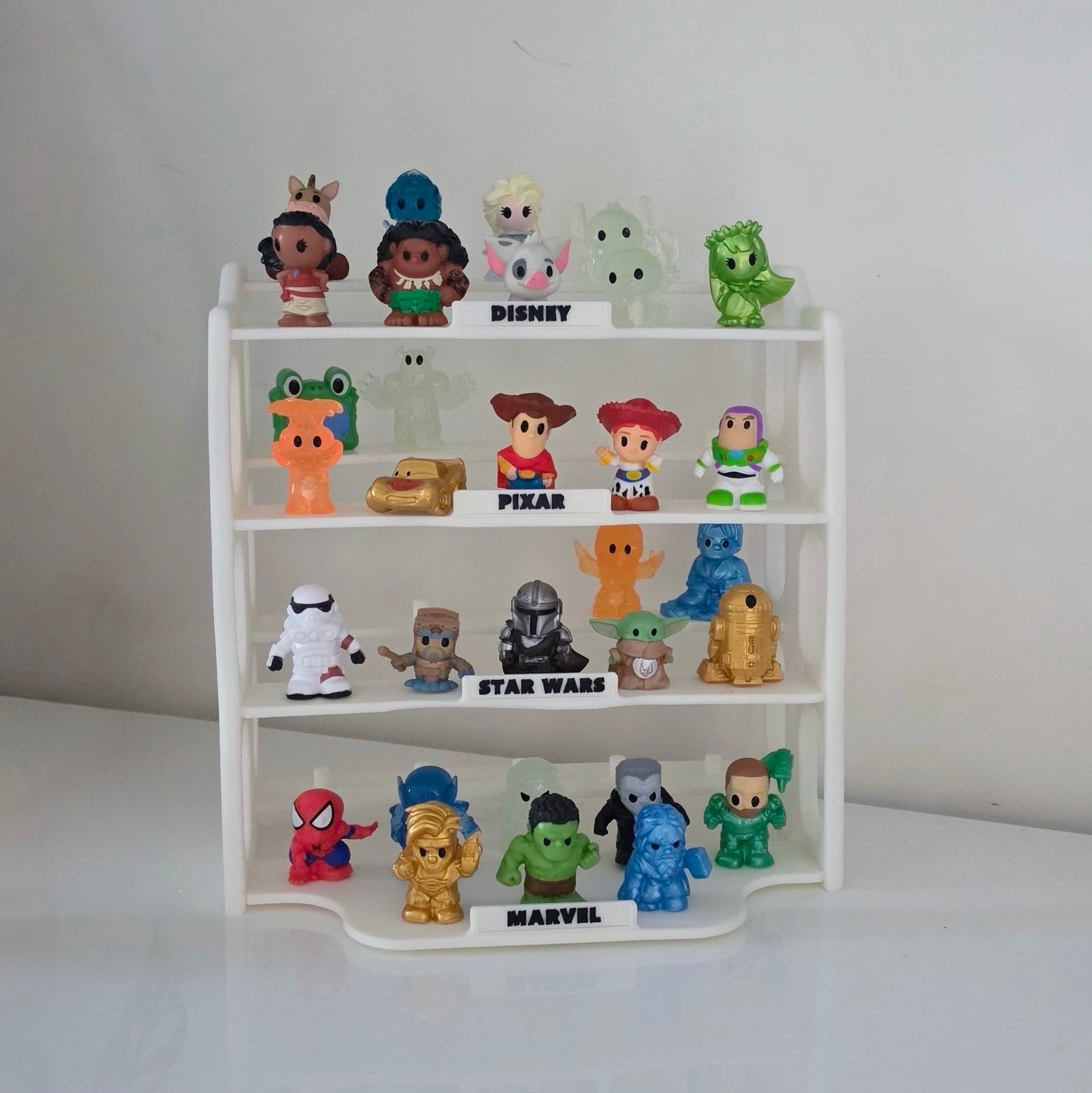 A white four-tier display stand holding small collectible character figures from Disney, Pixar, Star Wars, and Marvel, arranged on a white surface against a plain light background.