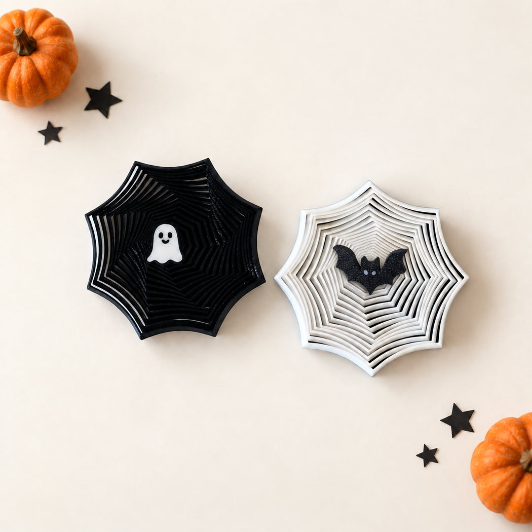 Two Halloween-themed spiderweb-shaped decorative pieces on a cream background, one black with a white ghost and one white with a black bat, surrounded by small orange pumpkins and black star cutouts.
