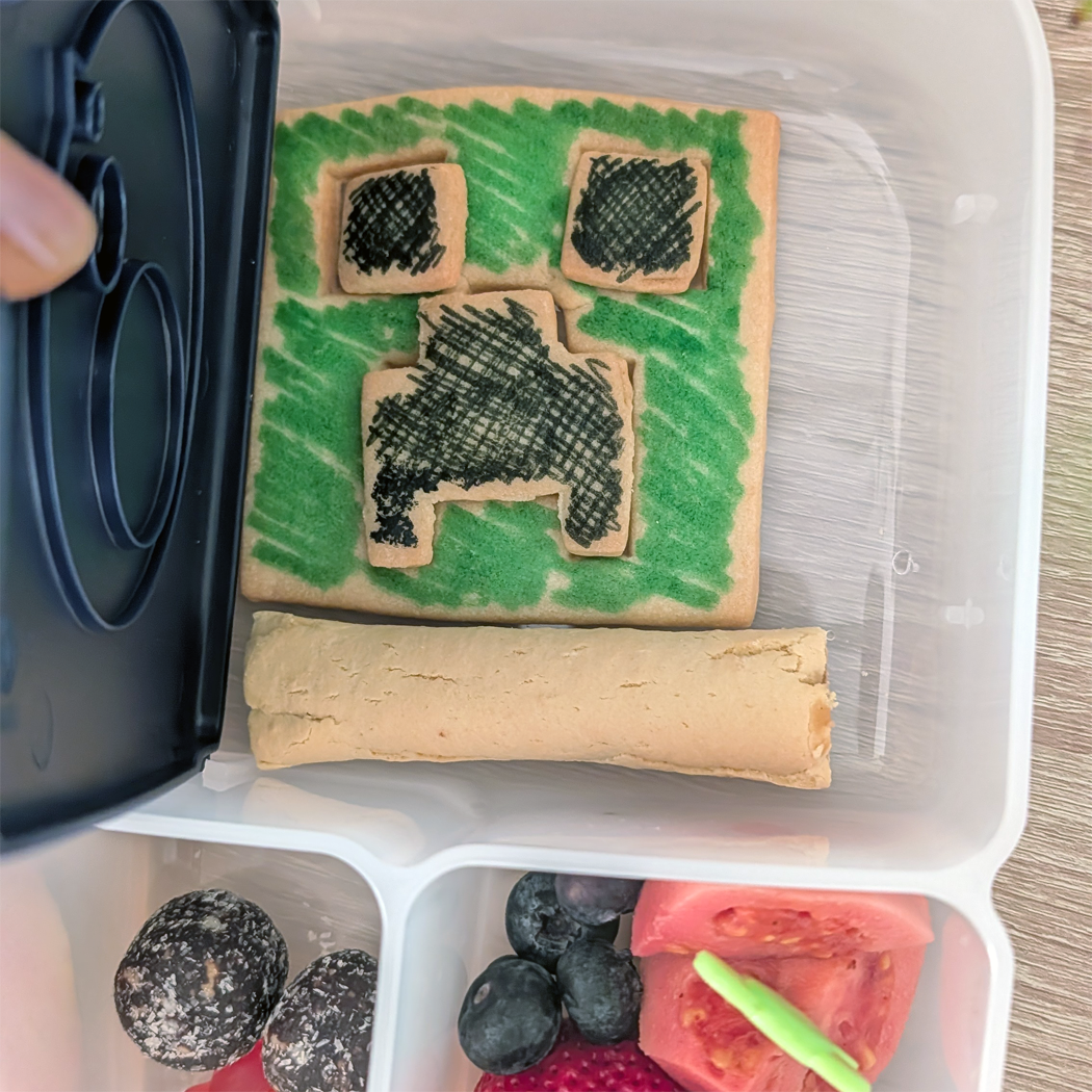 Bento box with a cookie shaped like a dinosaur, green icing, and other food items.