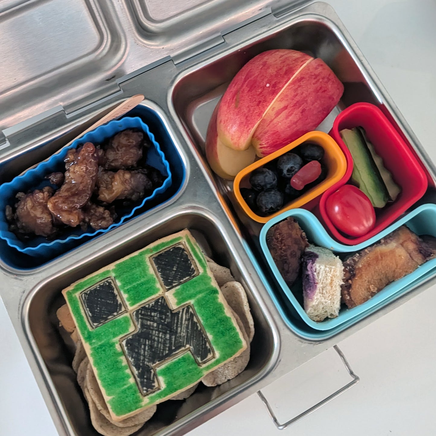 Children's lunchbox with compartments containing a cookie, fruit, and other snacks.