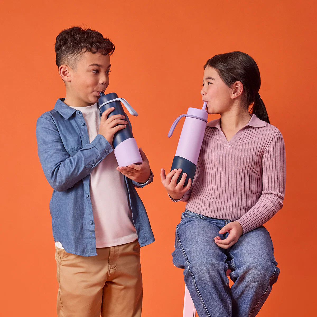 Two children holding and drinking from colorful water bottles against an orange background