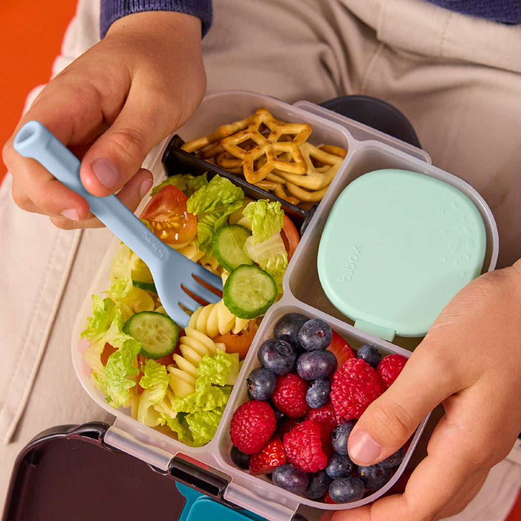 Child's hands holding a bento box with a variety of food including pasta salad, fruits, and vegetables.