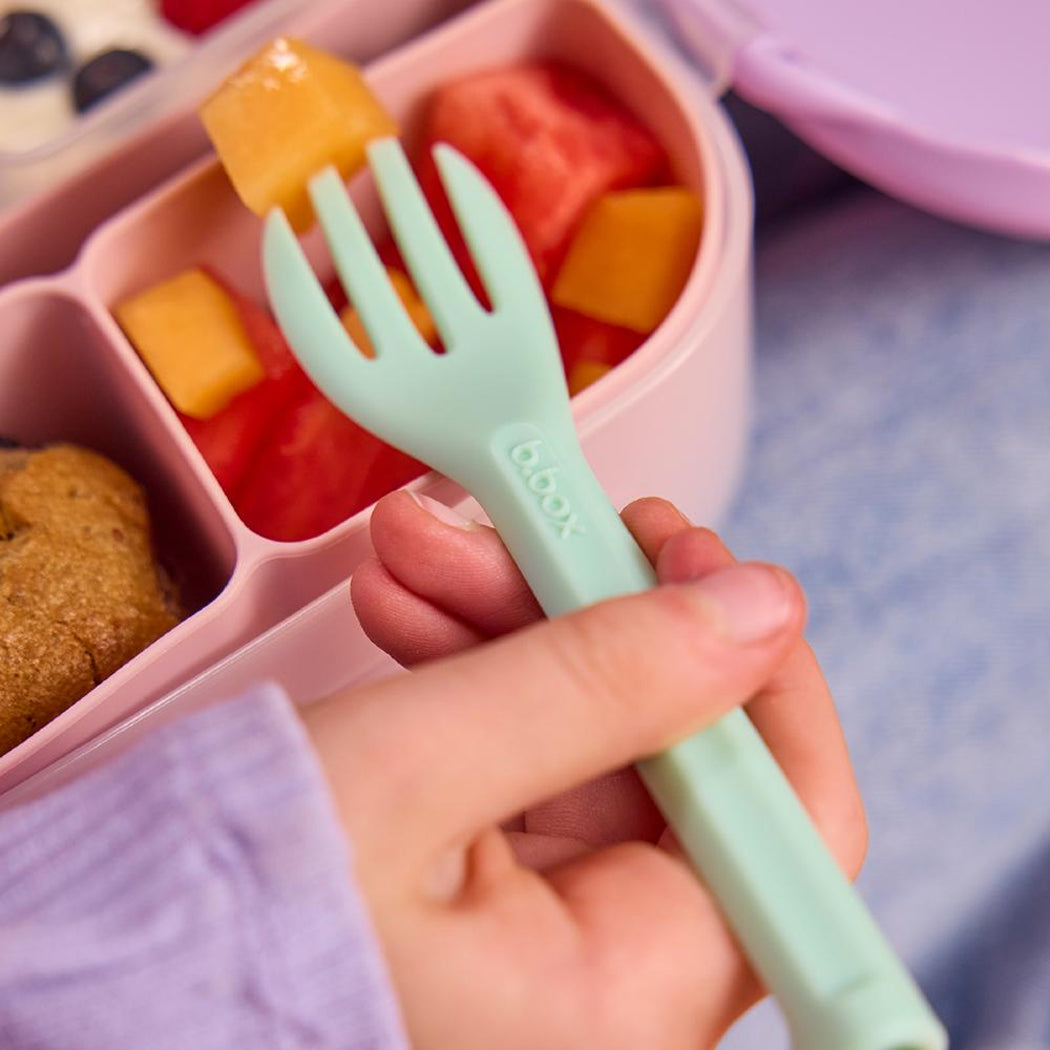 Child's hand holding a green fork over a pink compartmentalized food container with fruits.