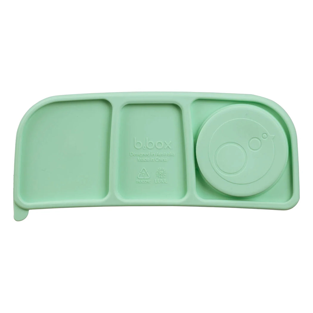 A mint green silicone baby feeding tray with three compartments and a round lid, shown on a plain white background. The brand name 'b.