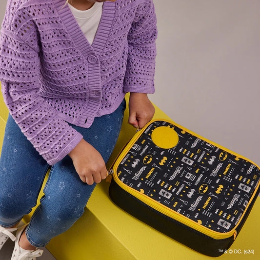 Child wearing a purple cardigan with a Batman-themed lunch bag on a yellow step.