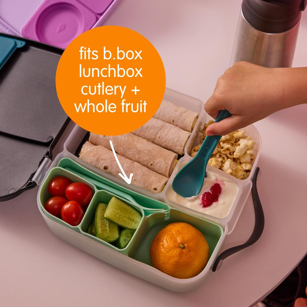 Bento box with food compartments including fruits and wraps, with text indicating compatibility with b.box lunchbox cutlery and whole fruit.