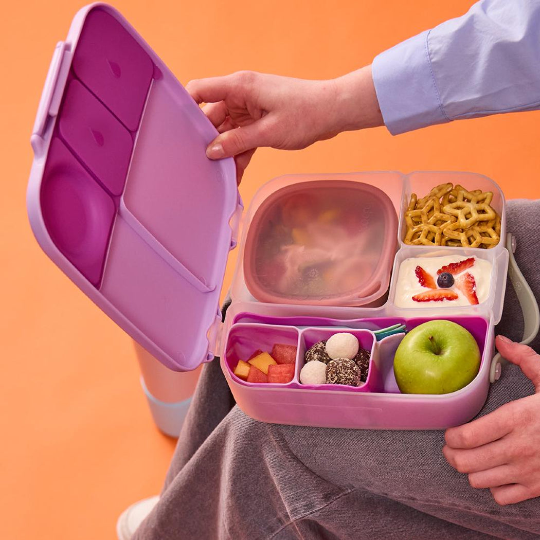 Person holding a pink bento box with compartments filled with food against an orange background