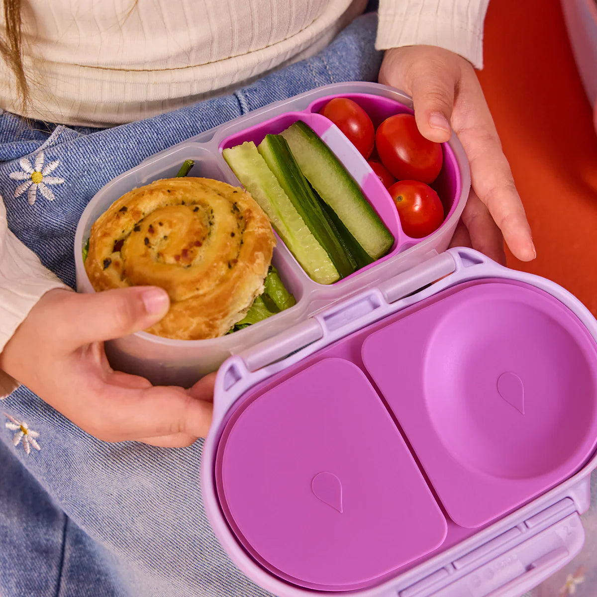 Purple bento box with compartments containing a pastry, cucumbers, and cherry tomatoes, held by a person.