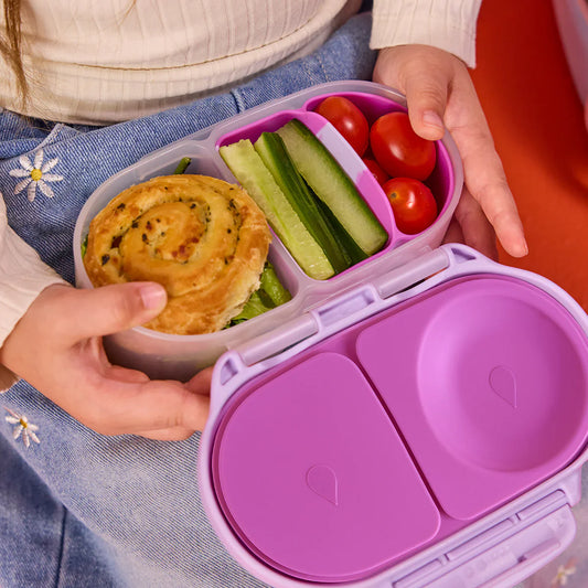 Purple bento box with compartments containing a pastry, cucumbers, and cherry tomatoes, held by a person.
