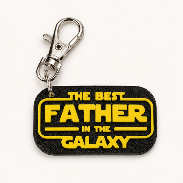 A black rectangular keychain with rounded corners featuring the yellow text 'THE BEST FATHER IN THE GALAXY' in a Star Wars-style font, attached to a silver metal lobster clasp, on a clean white background.