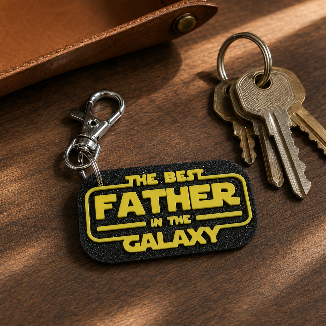 A black rectangular keychain with yellow text reading 'THE BEST FATHER IN THE GALAXY' is shown on a wooden surface next to metal keys and a brown leather strap.