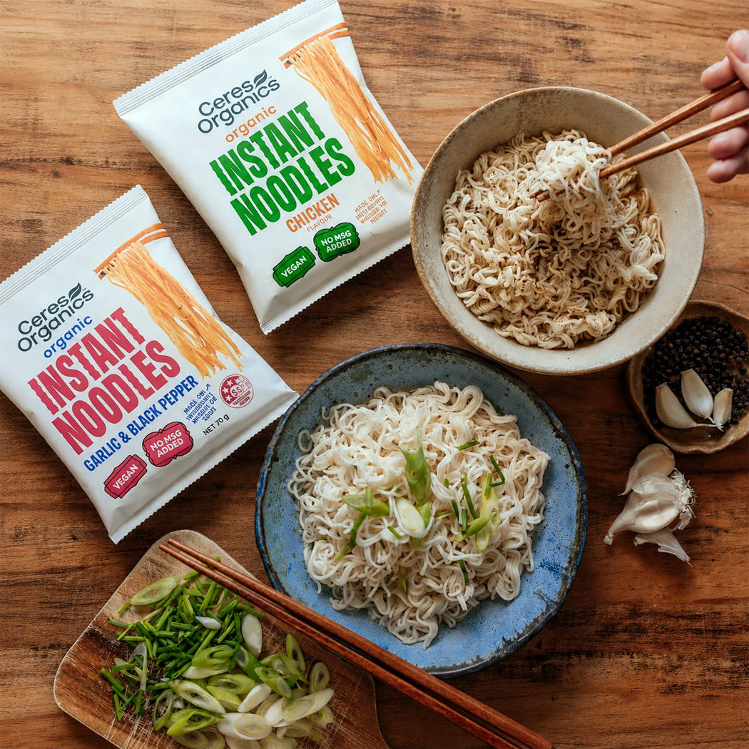 Two bags of Cerese Organics Instant Noodles, one Chicken and one Garlic & Black Pepper, arranged on a wooden table with bowls of prepared noodles, chopsticks, garlic, and green onions.