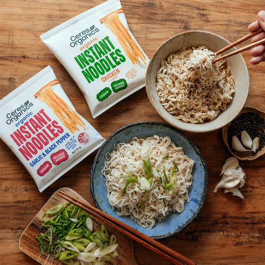 Two bags of Cerese Organics Instant Noodles, one Chicken and one Garlic & Black Pepper, arranged on a wooden table with bowls of prepared noodles, chopsticks, garlic, and green onions.