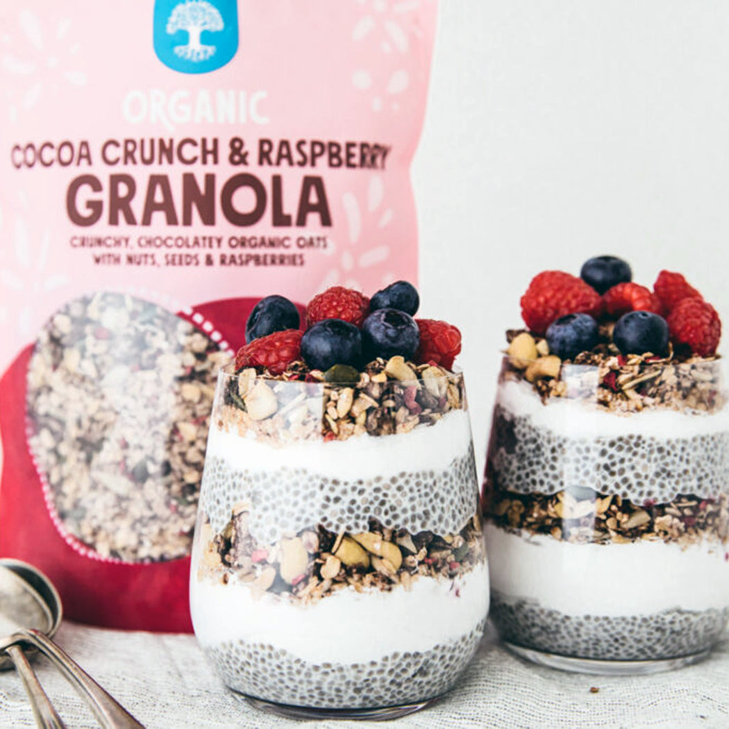 Two glasses of layered dessert with granola and berries next to a bag of Organic Cocoa Crunch & Raspberry Granola.