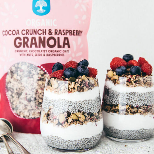 Two glasses of layered dessert with granola and berries next to a bag of Organic Cocoa Crunch & Raspberry Granola.