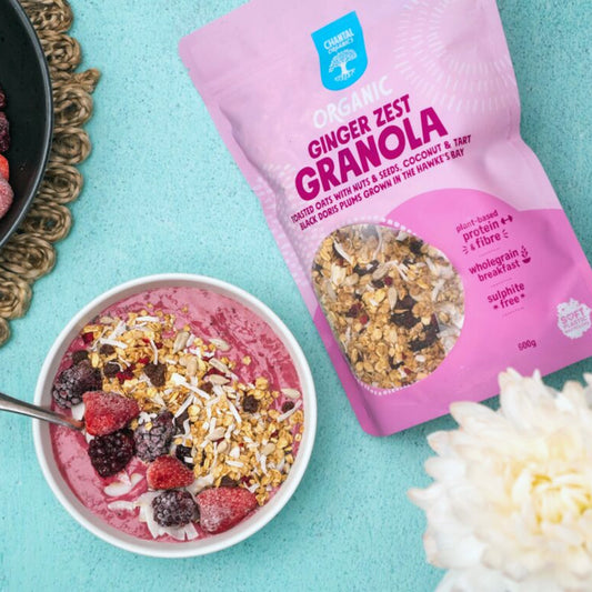 Bowl of granola with pink smoothie, ginger zest granola package, and white flower on a turquoise background