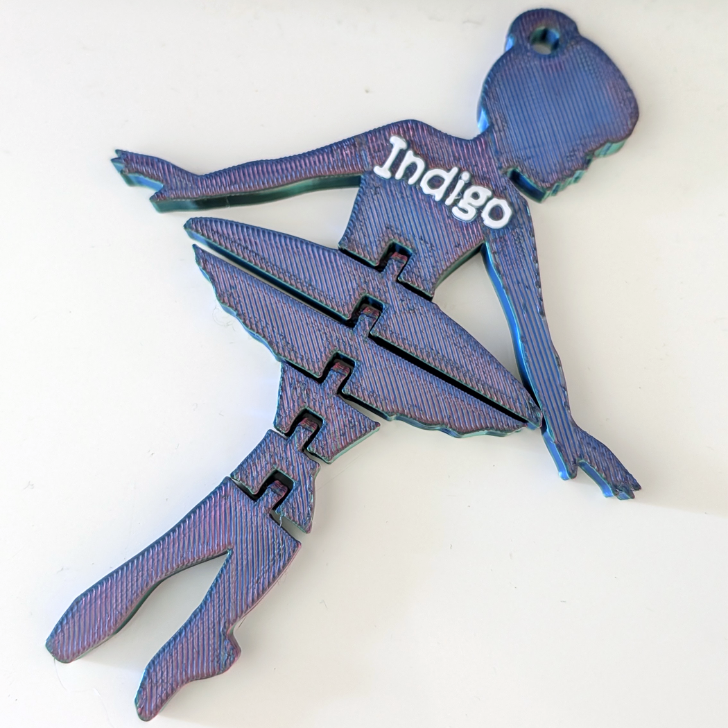 A 3D-printed ballerina-shaped ornament or puzzle piece in an iridescent indigo blue color, featuring the word 'Indigo' printed on its back, shown on a plain white background.
