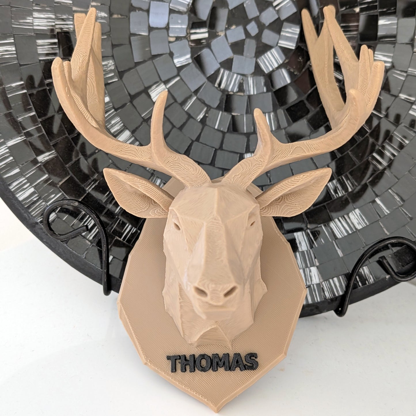 A personalized 3D-printed deer head wall mount in a light tan color, featuring the name 'THOMAS' in black lettering on the base. The mount is displayed in front of a black mosaic plate.