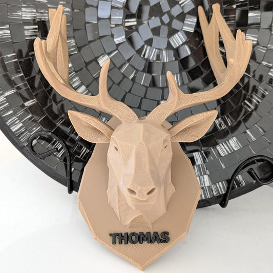 A personalized 3D-printed deer head wall mount in a light tan color, featuring the name 'THOMAS' in black lettering on the base. The mount is displayed in front of a black mosaic plate.