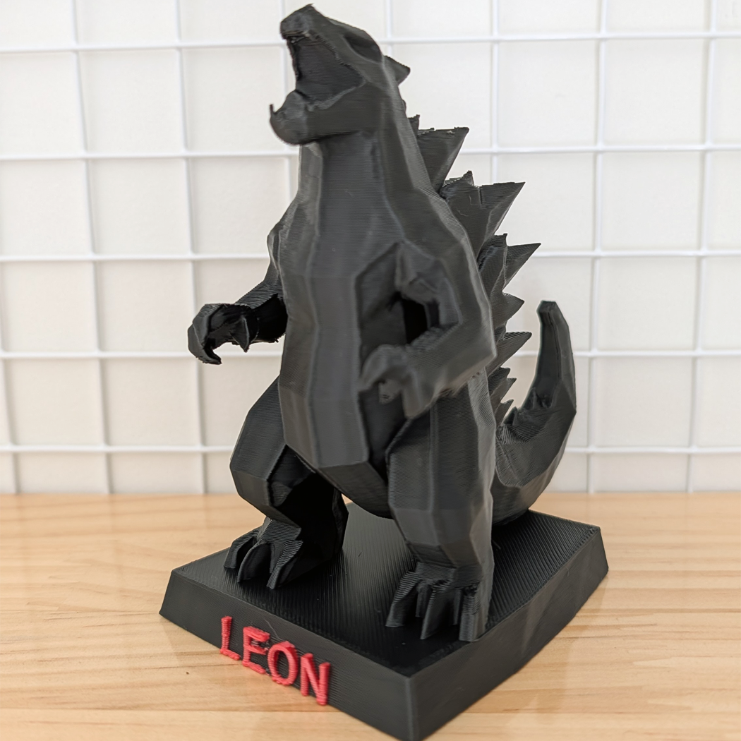 A black 3D-printed Godzilla figurine standing on a black base with the name 'LEON' in red letters, set on a wooden surface against a white grid background.