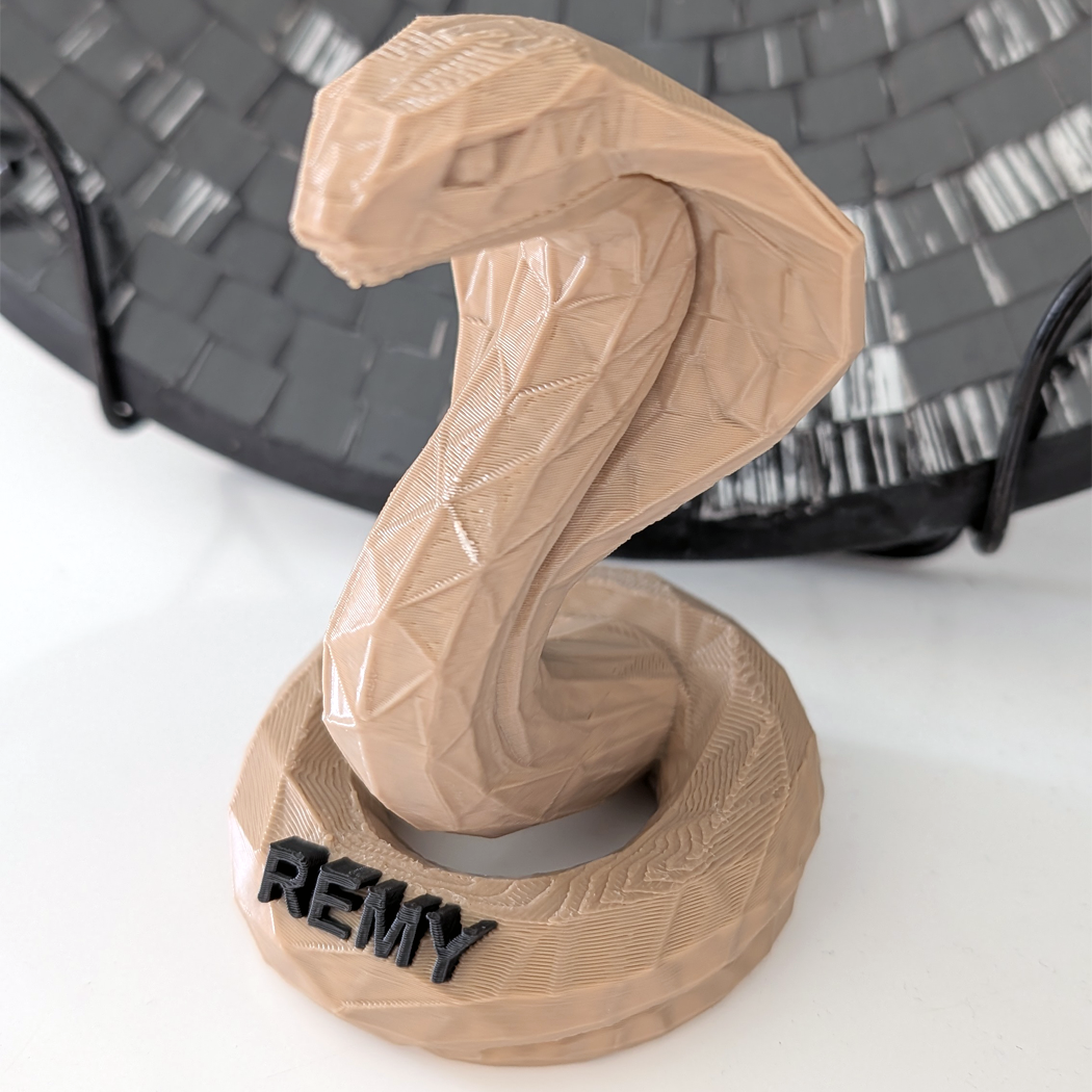 A 3D-printed cobra head sculpture on a matching circular base, with the name 'REMY' in black lettering on the front. The sculpture is a light tan/beige color, shown in a close-up shot against a white surface with a dark woven basket in the background.
